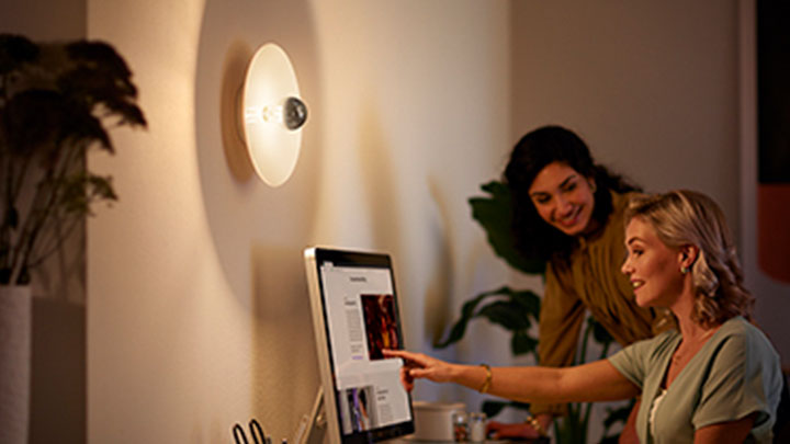Mujeres trabajando con un ordenador junto a una lámpara de espejo de LED de Philips de montaje en pared