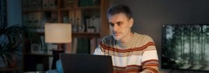 Un hombre con un jersey a rayas está sentado en su escritorio y mira el portátil que tiene delante