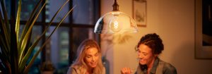 "Colgante de bombilla de filamento LED de estilo vintage que ilumina a dos mujeres que disfrutan de una conversación vespertina, creando un ambiente cálido en un apartamento urbano."