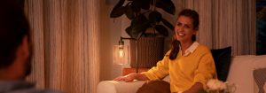 "Ambiente acogedor de sala de estar creado por una cálida lámpara de mesa LED, que ilumina a la mujer con suéter amarillo que disfruta de una conversación nocturna."