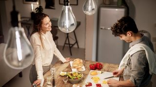 Una mujer observa a un hombre cortando verduras en una tabla de cortar en una cocina; se ven bombillas en la parte superior de la imagen